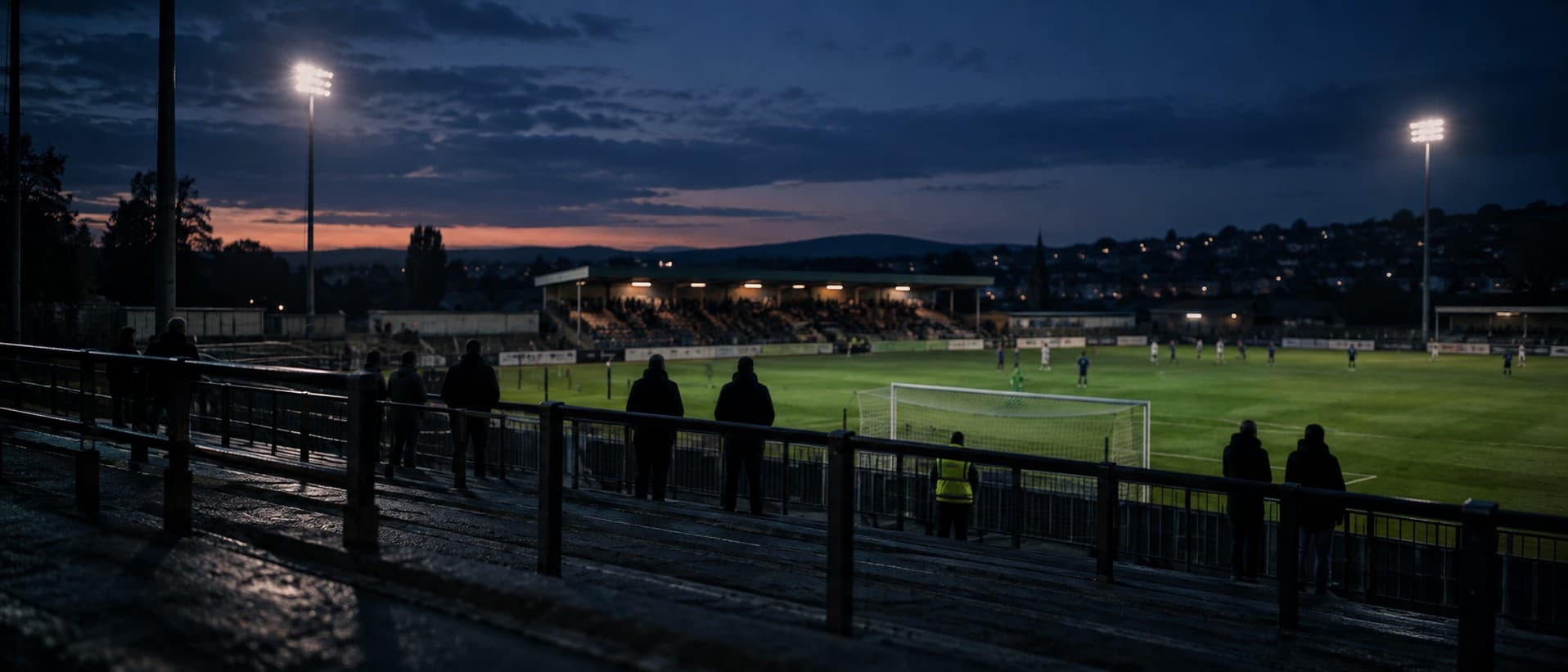 A small football ground at dusk, floodlights just on, supporters along the rail